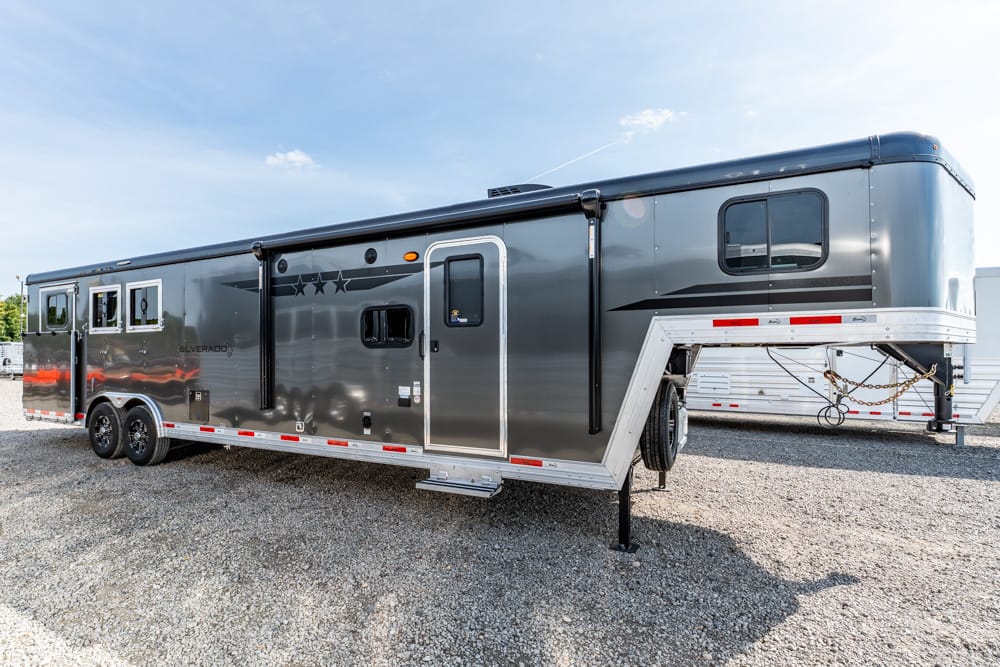 Gray gooseneck horse trailer with living quarters, parked on a gravel lot, side view showing windows and side door with steps.