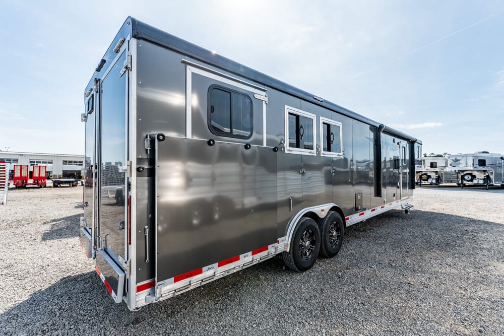 Long silver travel trailer with two axles and side windows parked on a gravel lot under a clear sky.