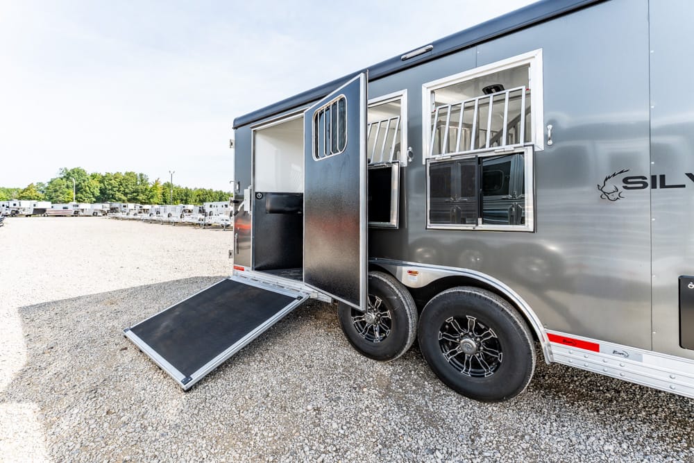 Gray horse trailer with a lowered ramp and open side door, exposing interior stalls and tack compartments on a gravel lot.