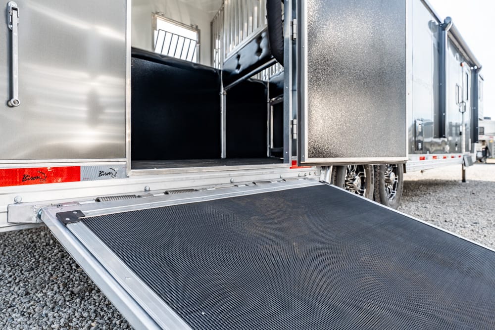 Aluminum trailer with its black ramp extended to ground, showing an empty, fenced-in interior and nearby wheels on gravel outside. It appears to be a cargo or livestock trailer ready for loading.