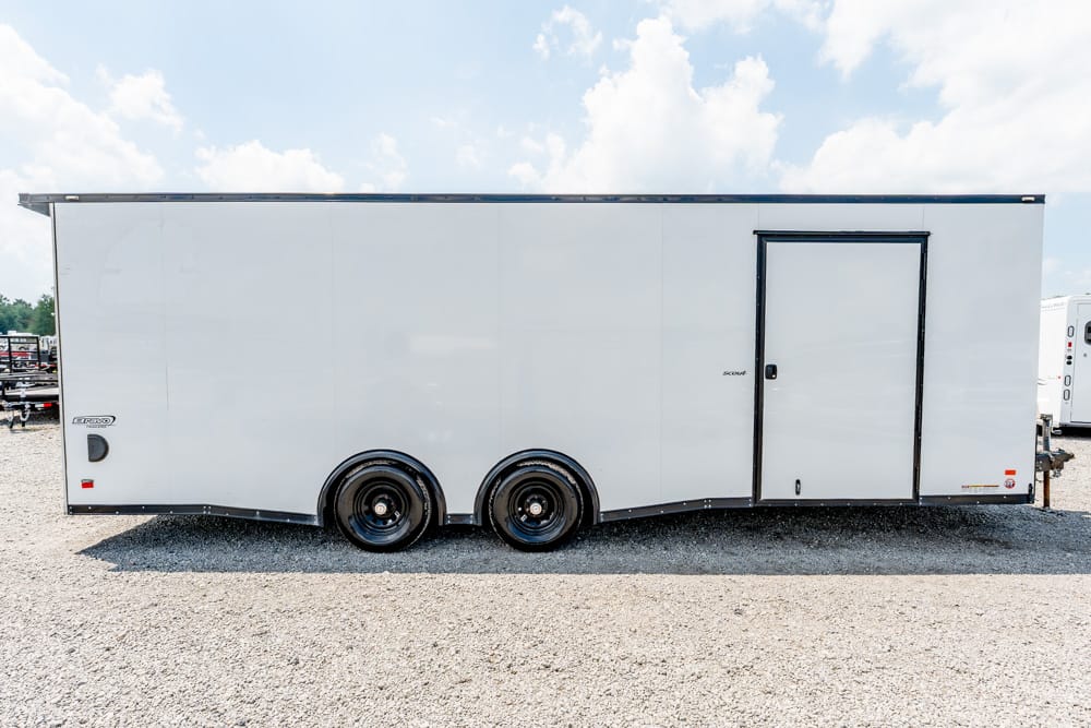 White enclosed cargo trailer with two axles parked on a gravel lot, featuring a side access door on the right.