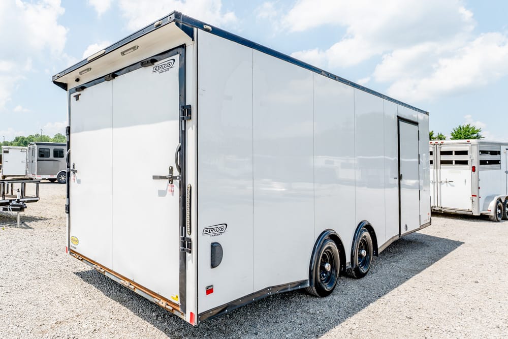 Side view of a large white enclosed cargo trailer with double rear doors and three axles, parked on a gravel lot.