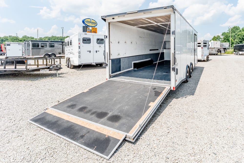 White enclosed trailer with rear loading ramp down, parked on a gravel lot with other trailers nearby.