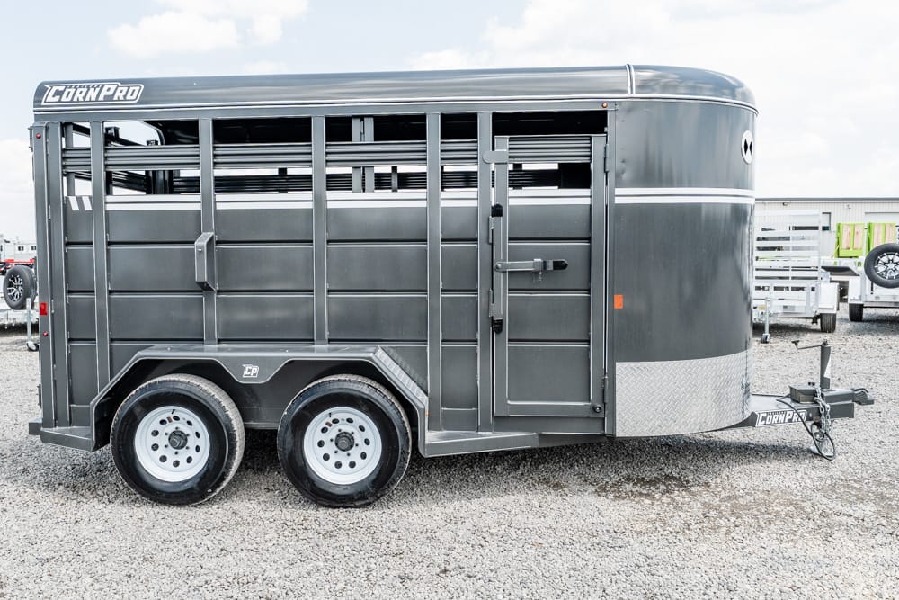 Grey CornPro livestock trailer with two axles and vented side panels on a gravel lot, hitch visible at the front.