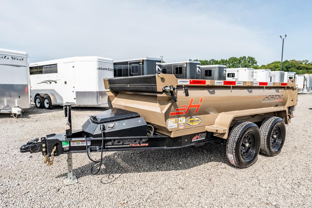 Tan Horizon dump trailer with dual axles and branding, hitched in a gravel lot with horse trailers in the background.