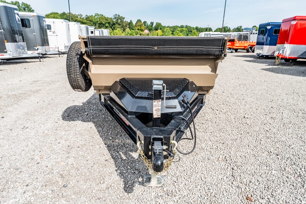 Front view of a black trailer hitch and beige dump bed with a spare tire on the side, parked on a gravel lot with other trailers in the background.