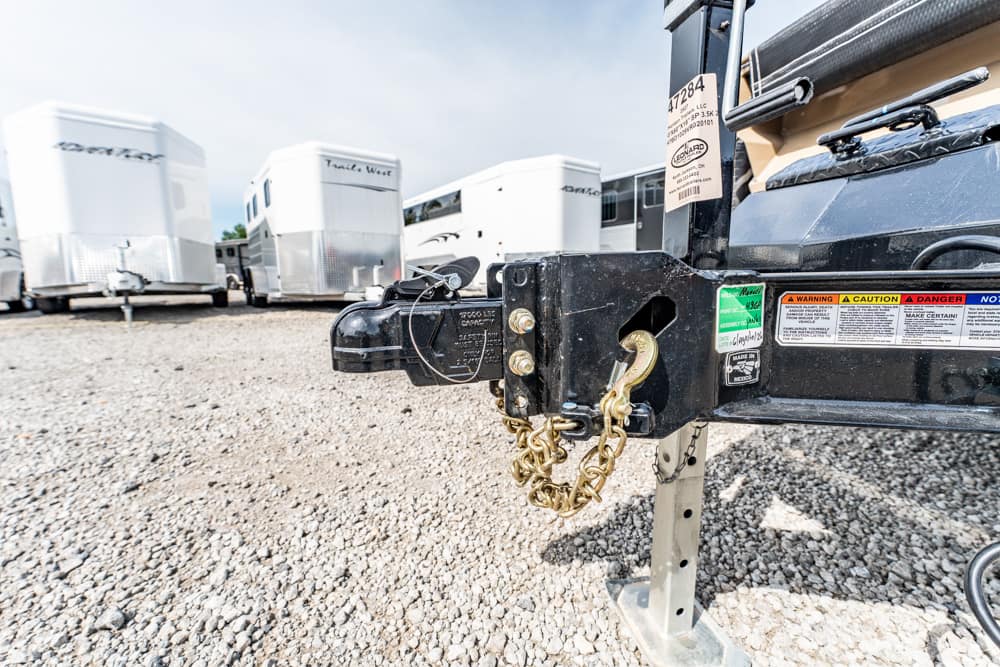 Close-up of a trailer hitch with safety chains and lock, on a gravel lot with white horse trailers in the background.