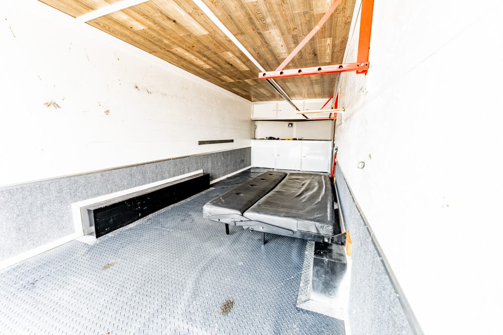 Interior of a small mobile workshop trailer: diamond-plate floor, white walls with gray lower panel, a black fold-out bed, and white cabinets at the far end under a red ceiling hoist track