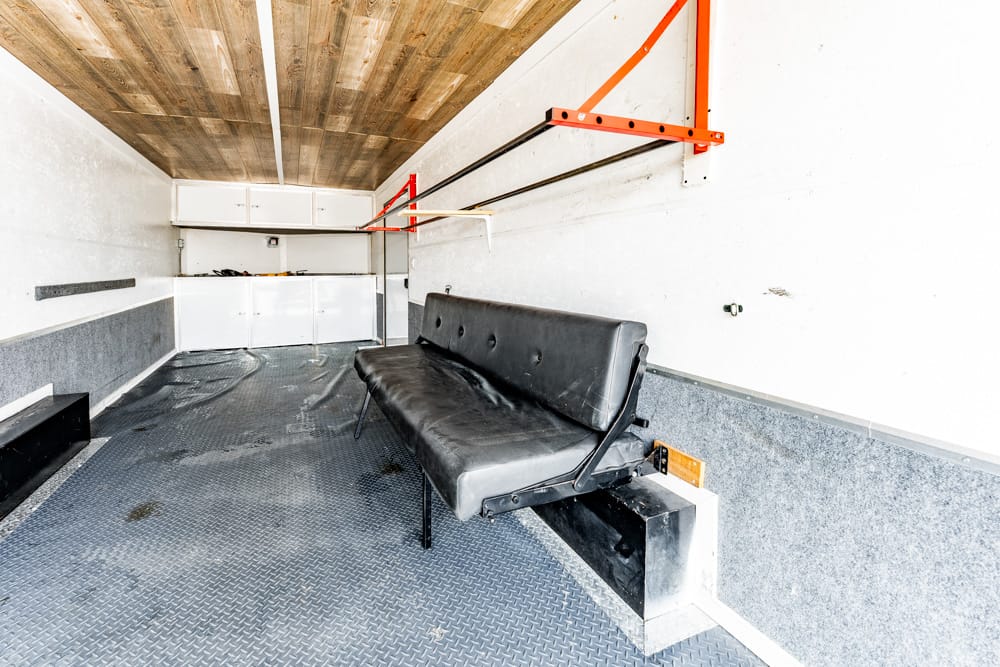 Interior of a small trailer/workshop with a black leather bench seat along the right wall, diamond-plate floor, and white storage cabinets at the far end.