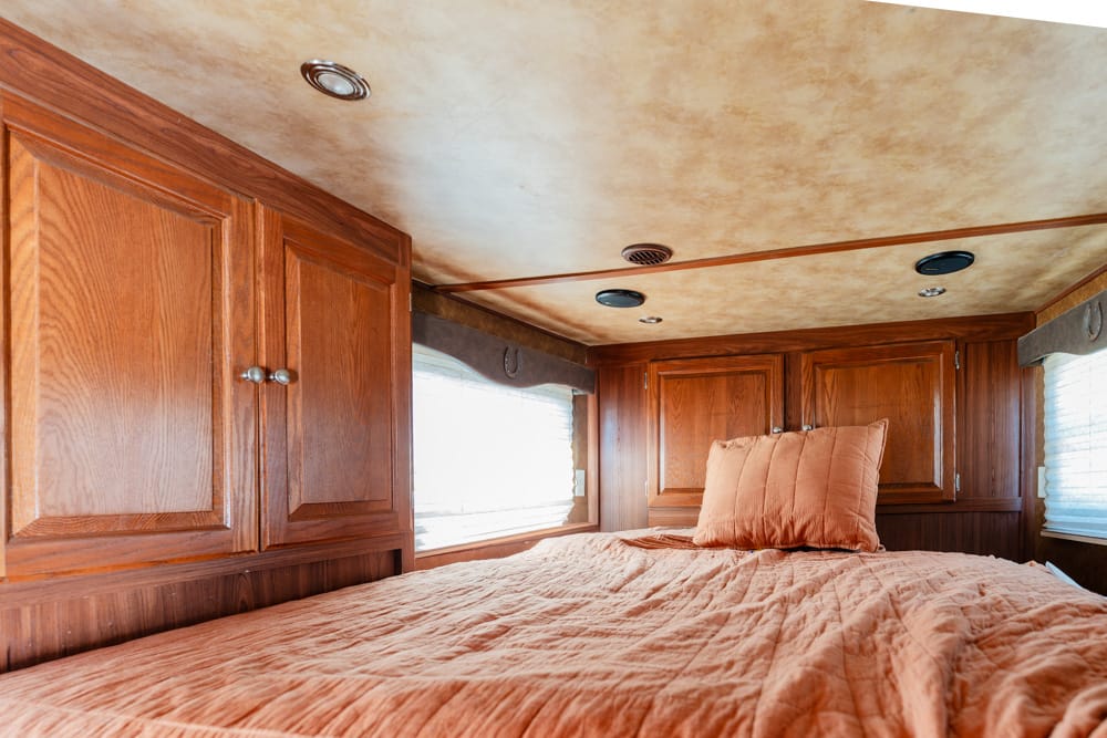 Wooden cabinets line the walls of a cozy RV bedroom, with an orange bedspread and pillow on a low bed.