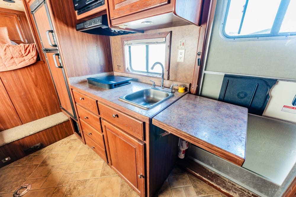 RV kitchen with wooden cabinets, stainless sink, and blue countertop; window over the sink of a camper interior.