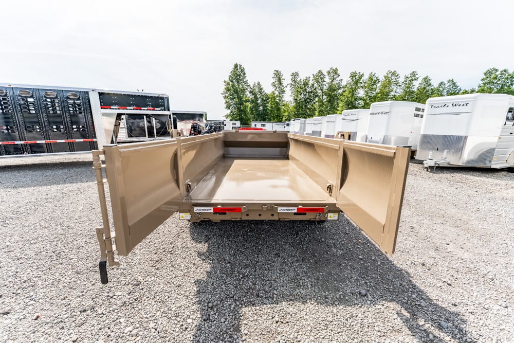 Beige dump trailer with rear doors open, in a gravel lot beside white horse trailers on a sunny day.