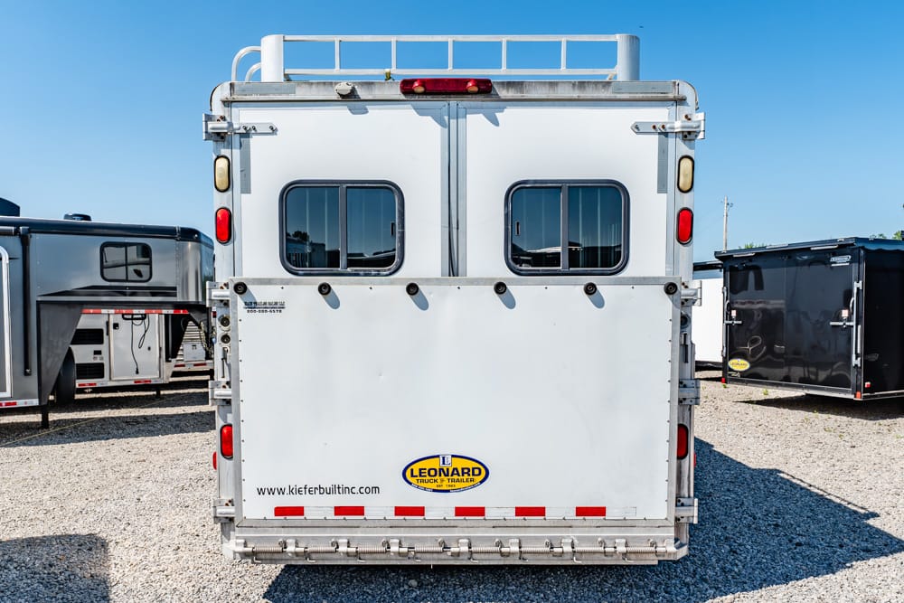 Rear view of a silver cargo trailer with two small windows and Leonard logo on the back door, on a gravel lot