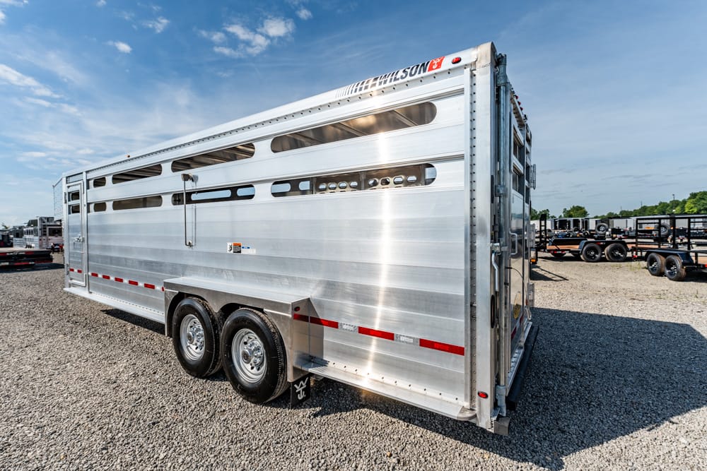 Polished aluminum two-axle livestock trailer with side vents on a gravel lot under a blue sky, Wilson branding visible at the top edge.