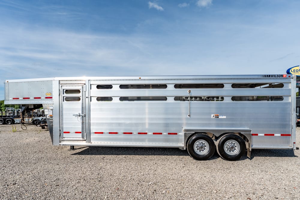 Silver livestock trailer with ventilation slats on a gravel lot under a blue sky, dual rear wheels visible on the side.