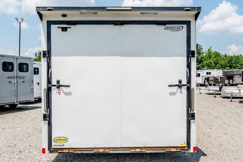 Back view of a white enclosed cargo trailer with double doors in a trailer yard; Bravo Trailers logo visible on the top right door edge.