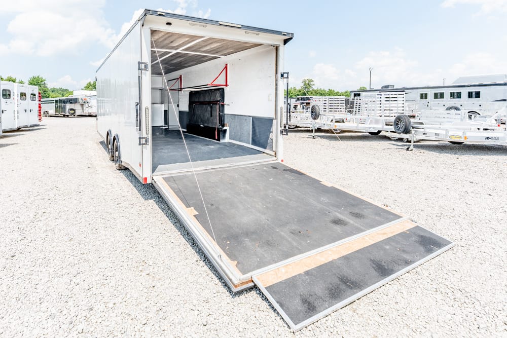 Open transport trailer with its loading ramp lowered, parked on a gravel lot with other trailers in the background.
