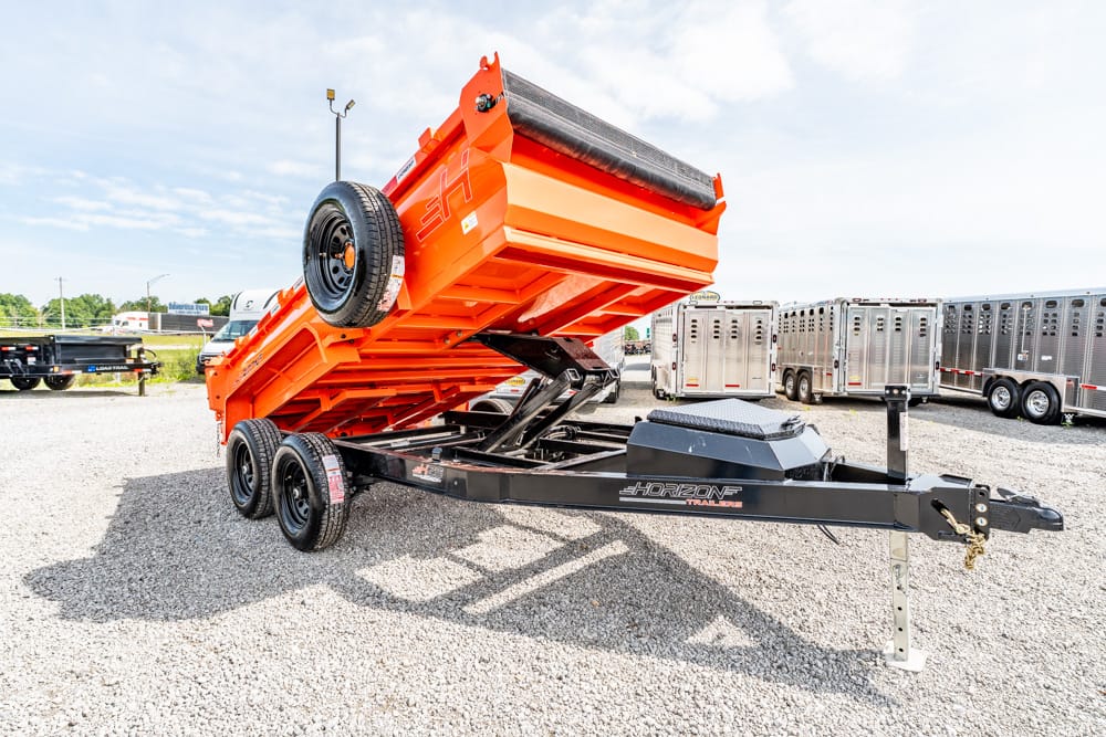 Orange tilt-bed trailer on a gravel lot with a spare wheel, raised for unloading, other trailers in the background