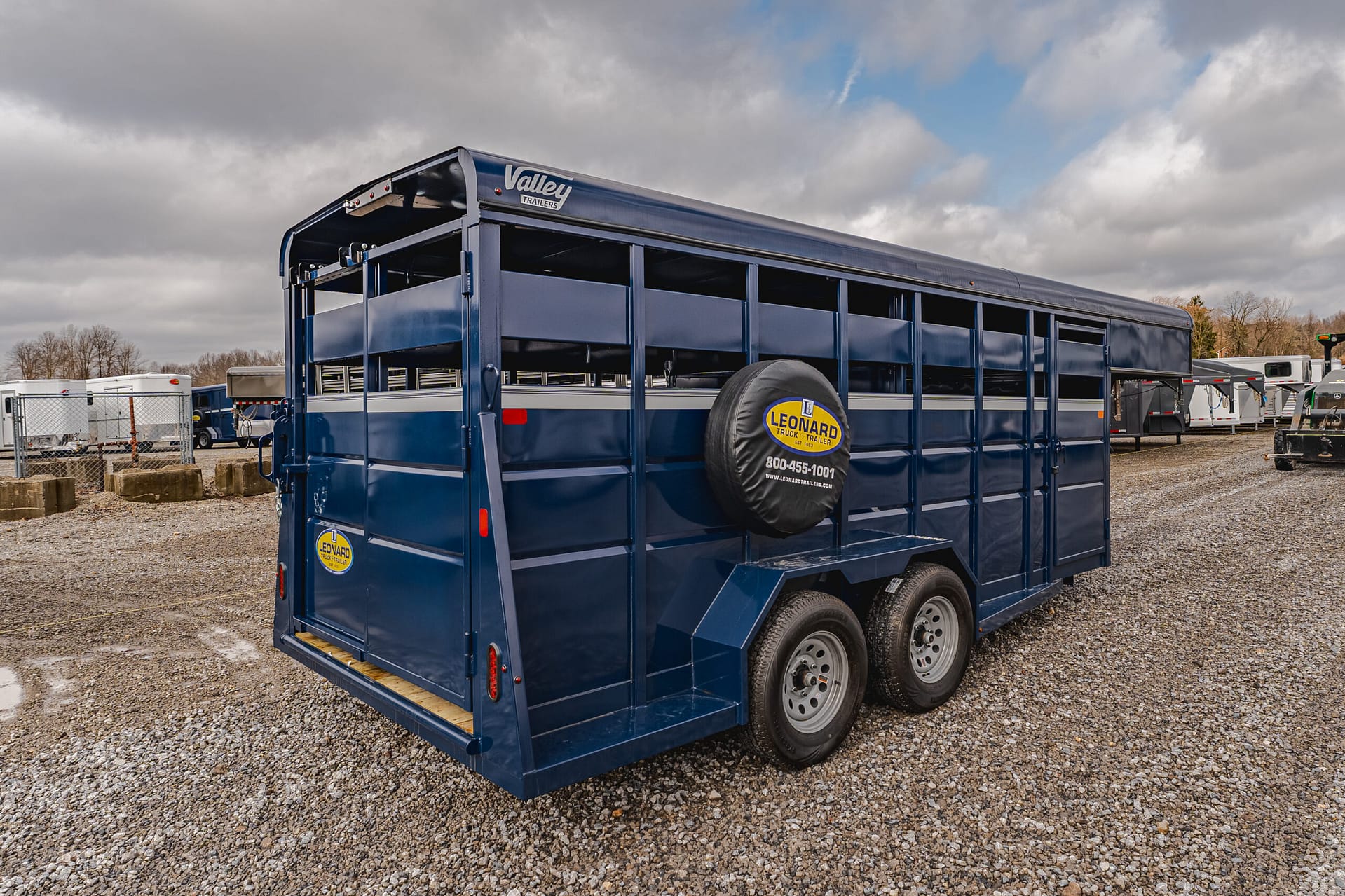 Blue livestock trailer with a Leonard Truck & Trailer spare tire cover, parked in a gravel lot with other trailers under a cloudy sky.