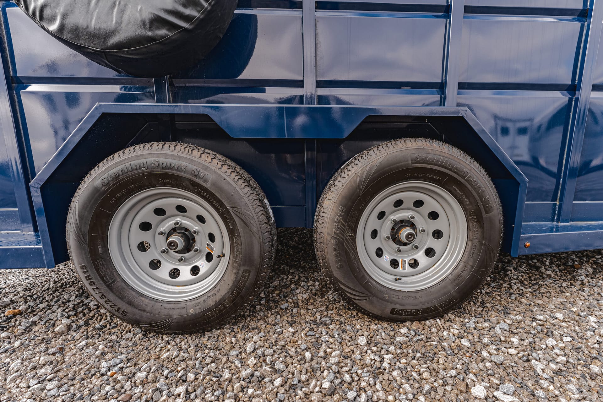 Two blue trailer wheels with metal fenders on a gravel surface, showing a mounted spare tire at the top-left edge of the trailer.