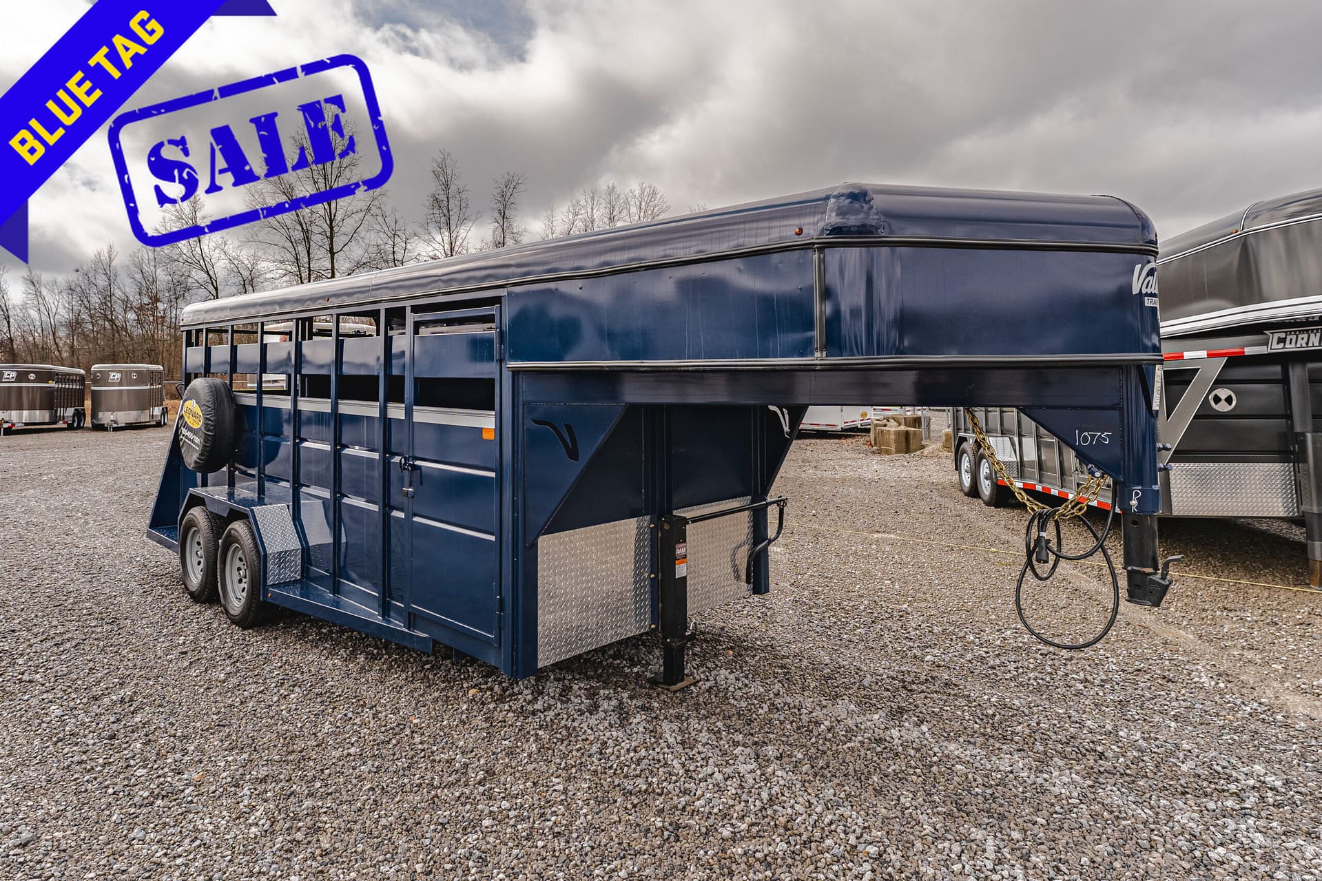 Blue cattle trailer on a gravel lot with a large blue SALE stamp and a diagonal BLUE TAG ribbon in the corner.