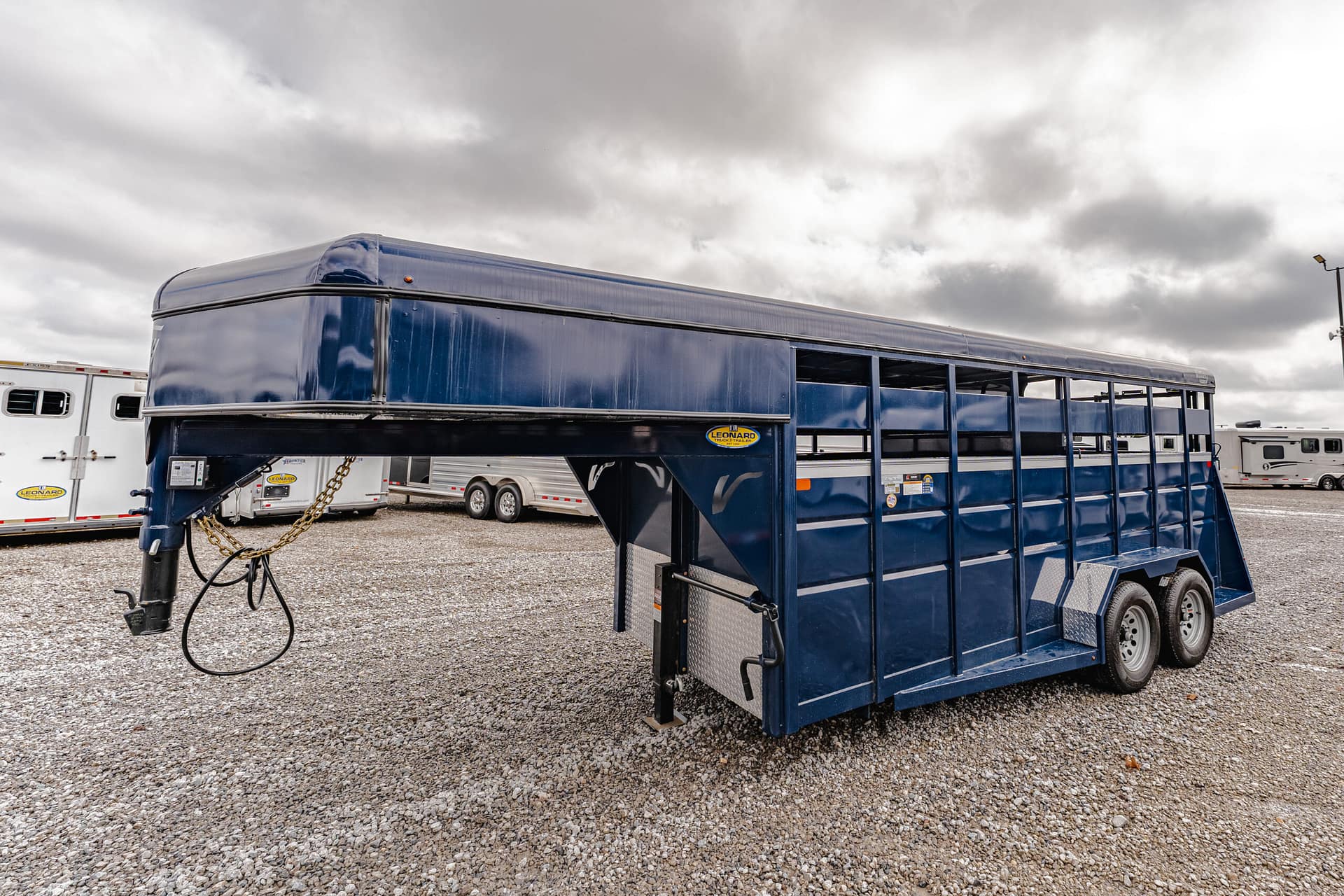 Blue livestock trailer with open side panels and dual axles sits on a gravel lot, with other trailers in the background under a cloudy sky.