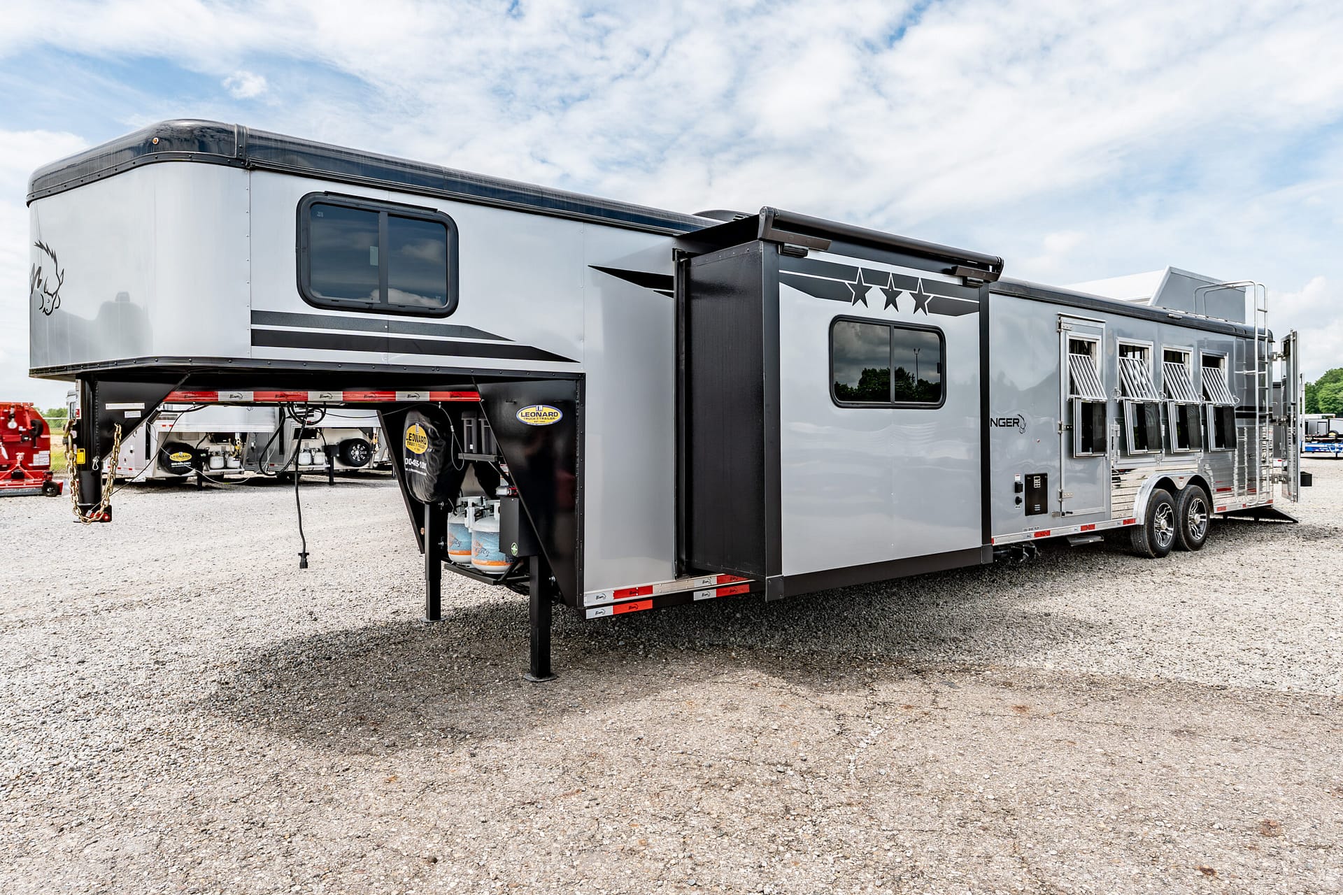 Gray and black gooseneck horse trailer with side windows and star graphics, parked on a gravel lot under a partly cloudy sky.