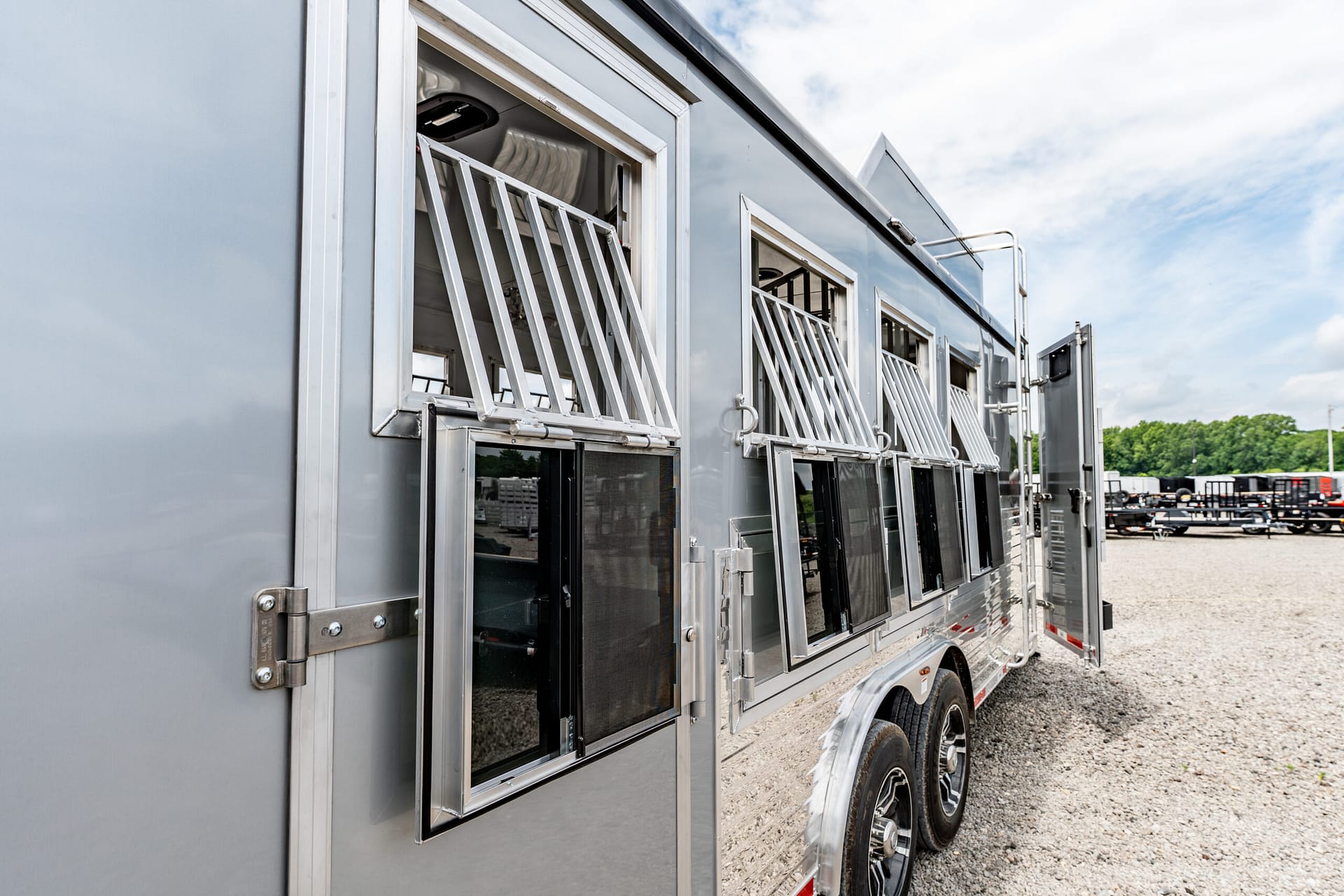 Side of a gray livestock trailer with open ventilated windows along the length, parked on a gravel lot under a cloudy sky