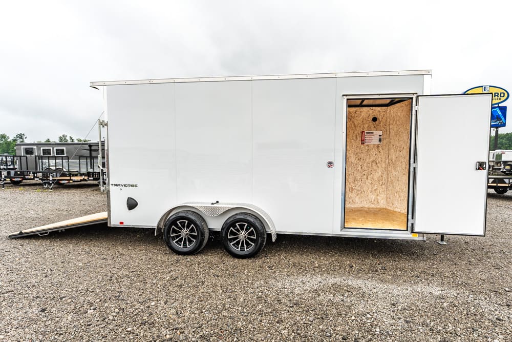 White enclosed cargo trailer with two axles on gravel, rear door open and ramp down, exposing unfinished wooden interior and plywood walls on a trailer lot.