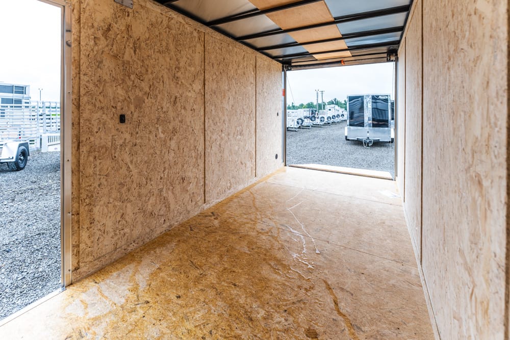 Empty plywood-walled trailer interior with open back showing a gravel lot and parked trailers outside