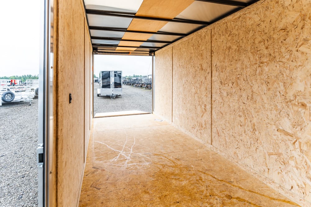 Interior of an empty plywood trailer, looking toward the rear opening with a gravel lot and trailers outside