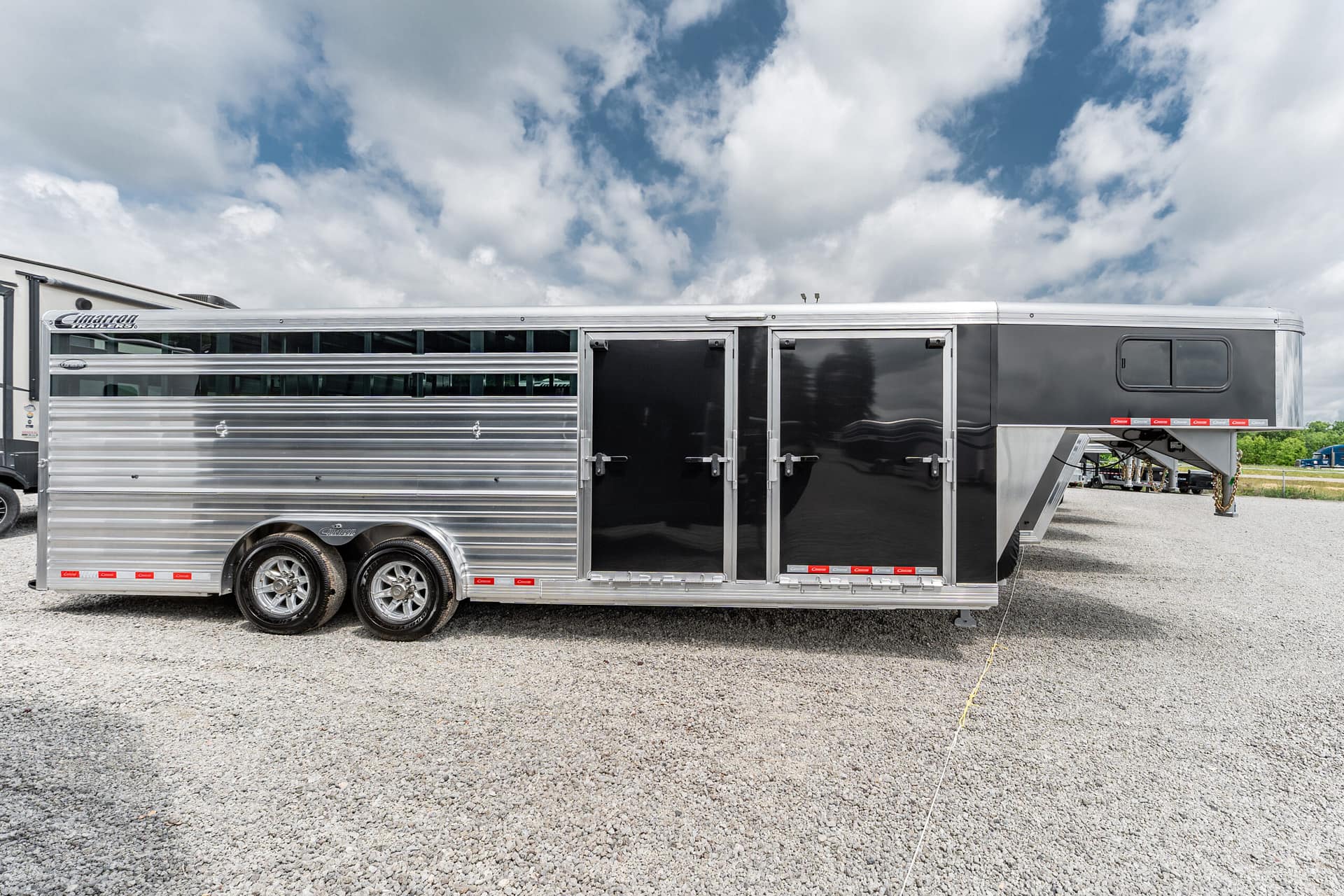 Silver-and-black horse trailer parked on a gravel lot beneath a cloudy sky, showing locking doors and vented side windows