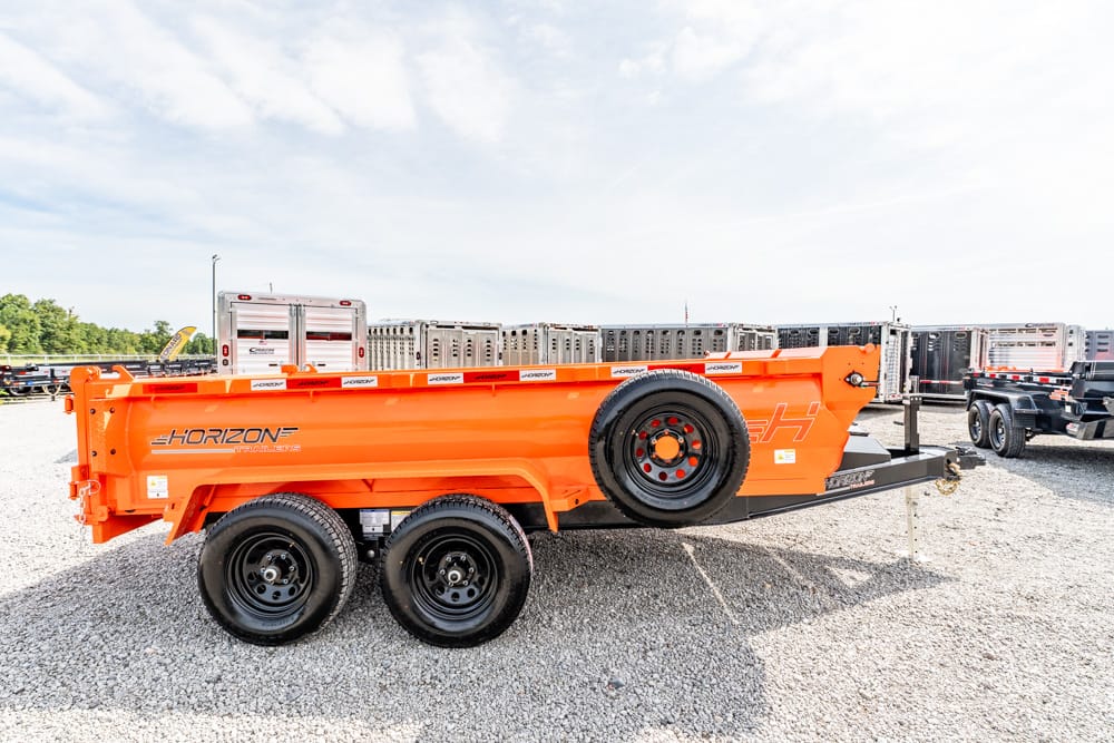 Orange Horizon trailer with a spare tire mounted on the side, twin axles, and hitch, parked in a gravel trailer lot with other trailers nearby.