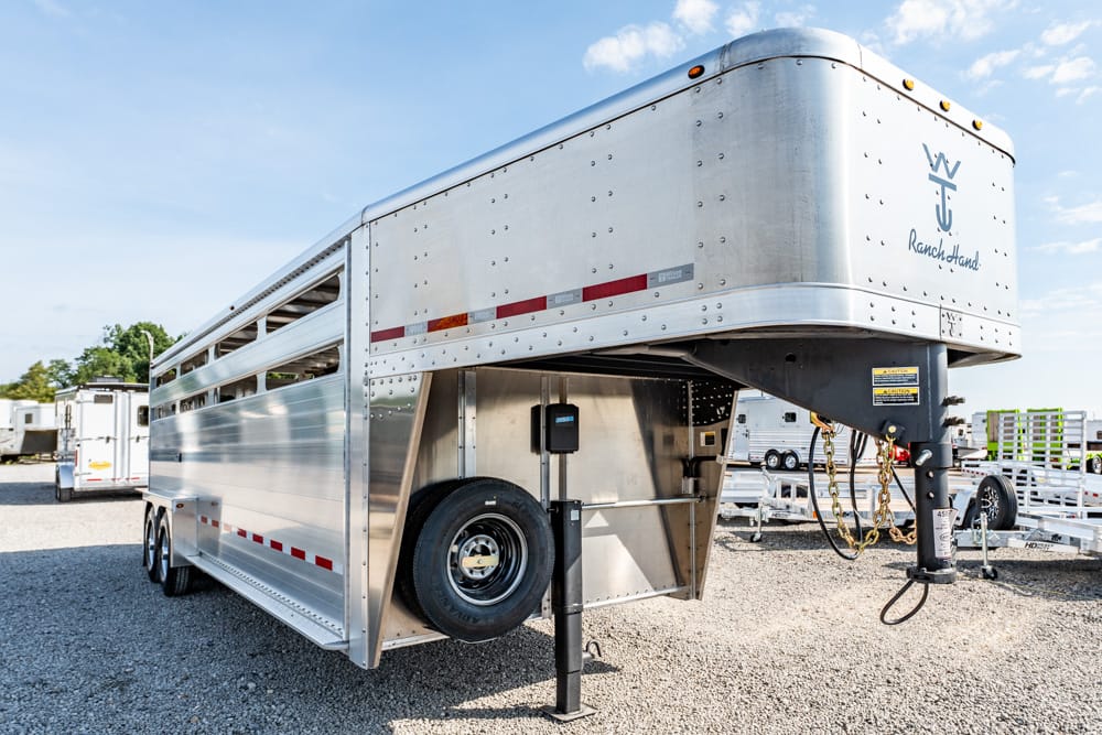 Silver gooseneck cattle trailer parked on gravel, front hitch with safety chains and spare tire inside nearby.] ,