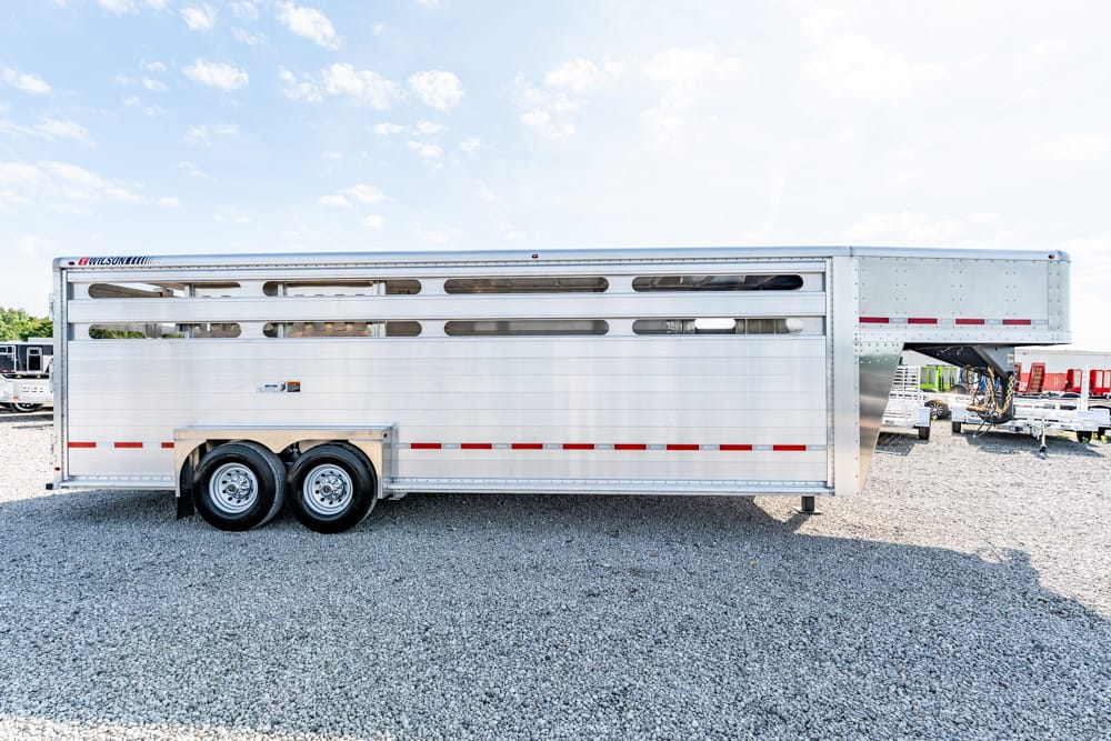 Silver aluminum livestock trailer with ventilation slots and dual wheels parked on a gravel lot under a blue sky after a sunny day.