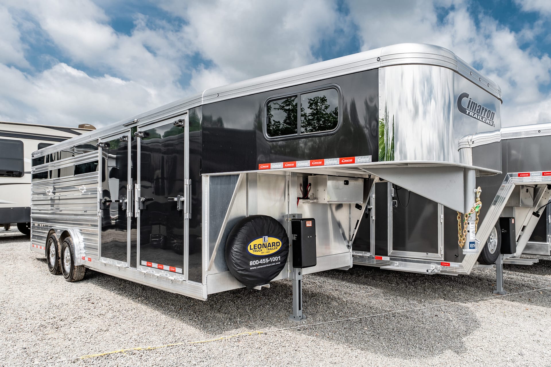 Side view of a large Cimarron horse trailer with black and silver panels, spare tire cover reading Leonard Truck & Trailer, parked on gravel under a blue sky with clouds.