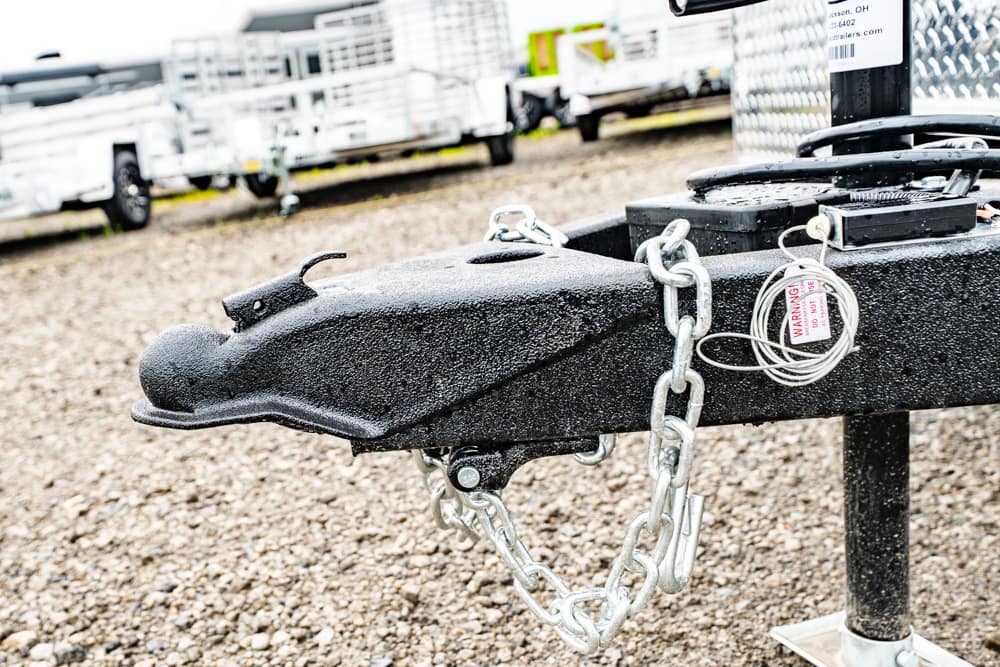 Close-up of a black trailer hitch with a metal chain and padlock, on a gravel lot with other trailers in the background.