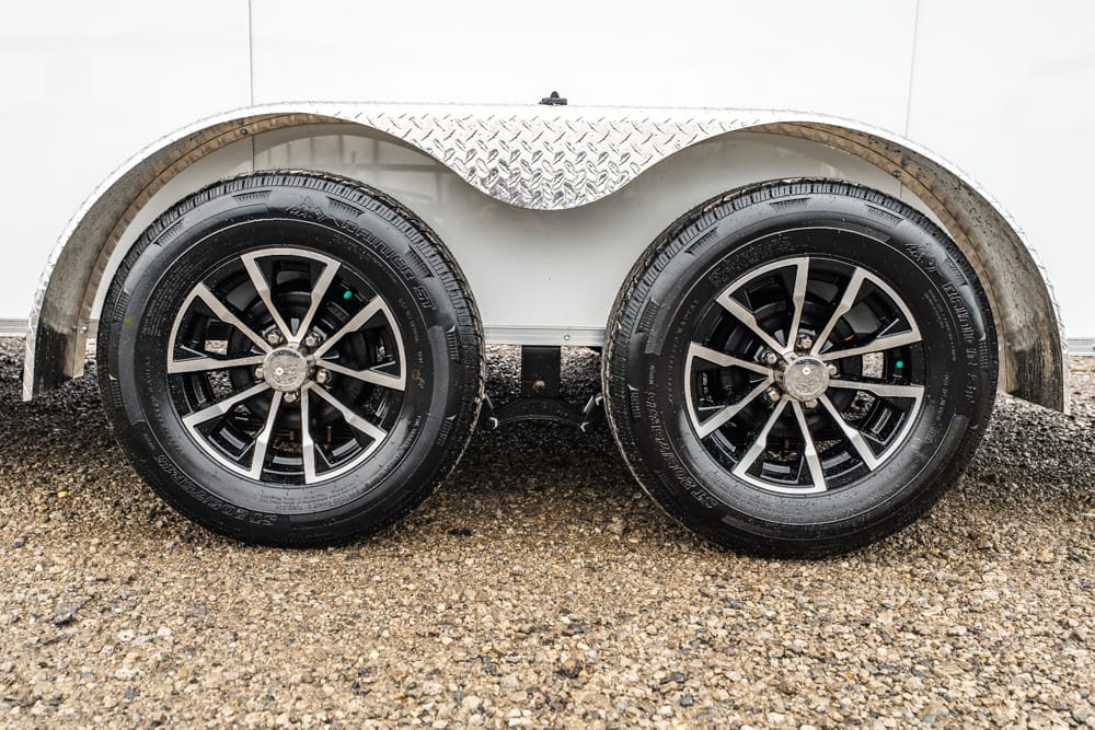 Dual trailer wheels with silver spoke rims on a gravel lot, undercarriage and diamond-plate wheel well visible.