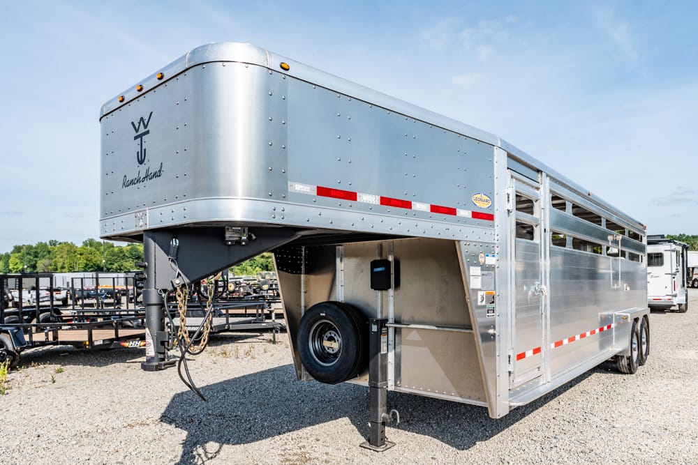 Silver gooseneck livestock trailer with spare tire mounted on the front, parked on gravel.