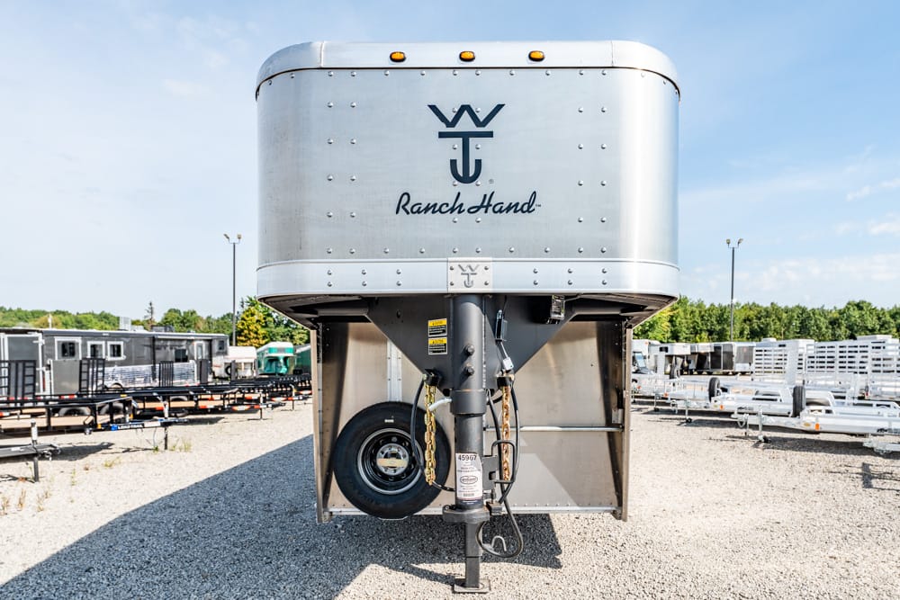 Front view of a silver Ranch Hand gooseneck trailer in a gravel lot with other trailers in the background under a blue sky.
