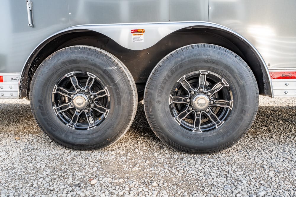 Double trailer wheels with black and chrome rims on a gravel surface beside a silver trailer body.