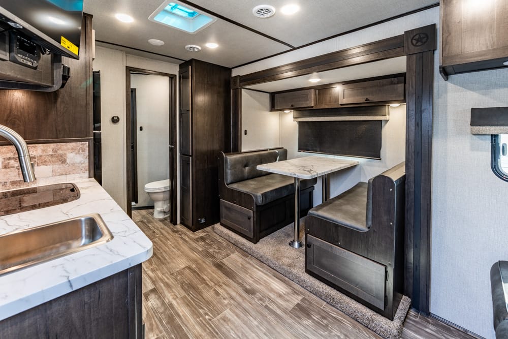 RV kitchen and dinette area with dark wood cabinets, marble countertop, and a booth-style dining table with seating on both sides