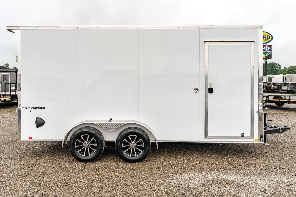 White enclosed trailer with a side door and dual-axle diamond-plate fenders on a gravel lot.