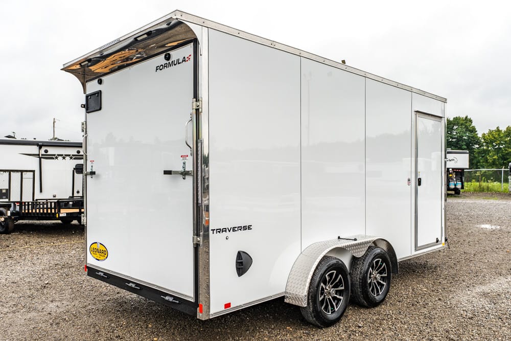 White enclosed cargo trailer with twin axles and diamond-plate fenders parked on a gravel lot, viewed from rear-left.