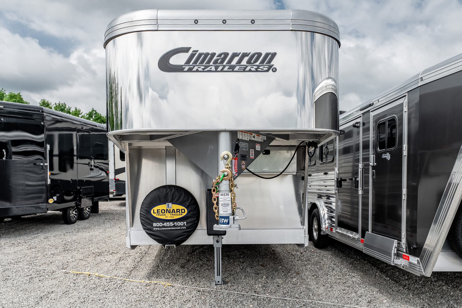 Shiny silver Cimarron Trailers trailer hitch with chains and spare tire cover in foreground, other trailers parked nearby under a cloudy sky