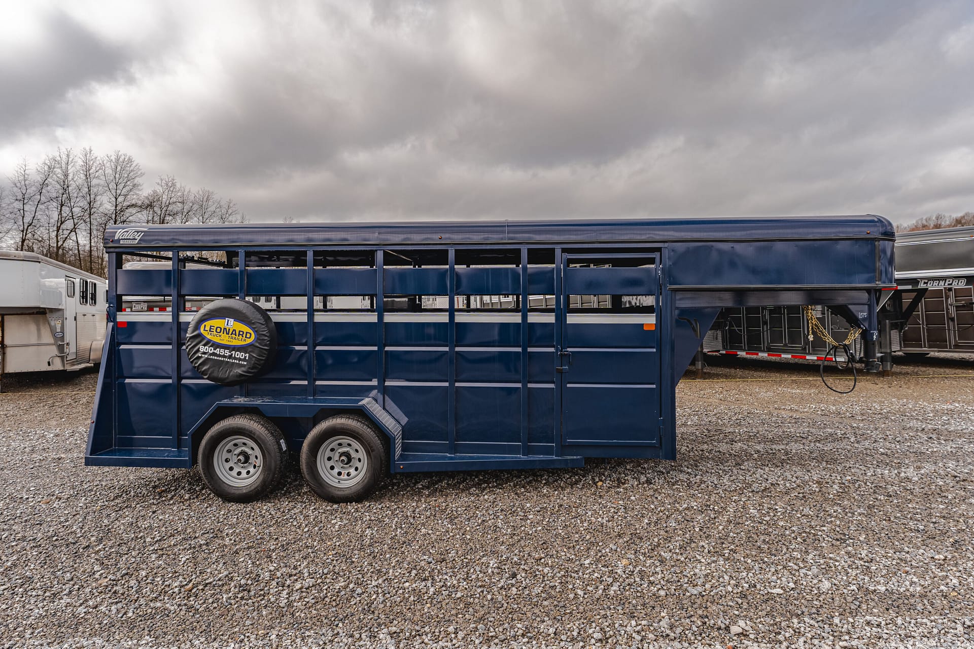 Blue livestock trailer on a gravel lot under a cloudy sky, with a spare tire cover labeled 'Leonard Trailer' visible on the side