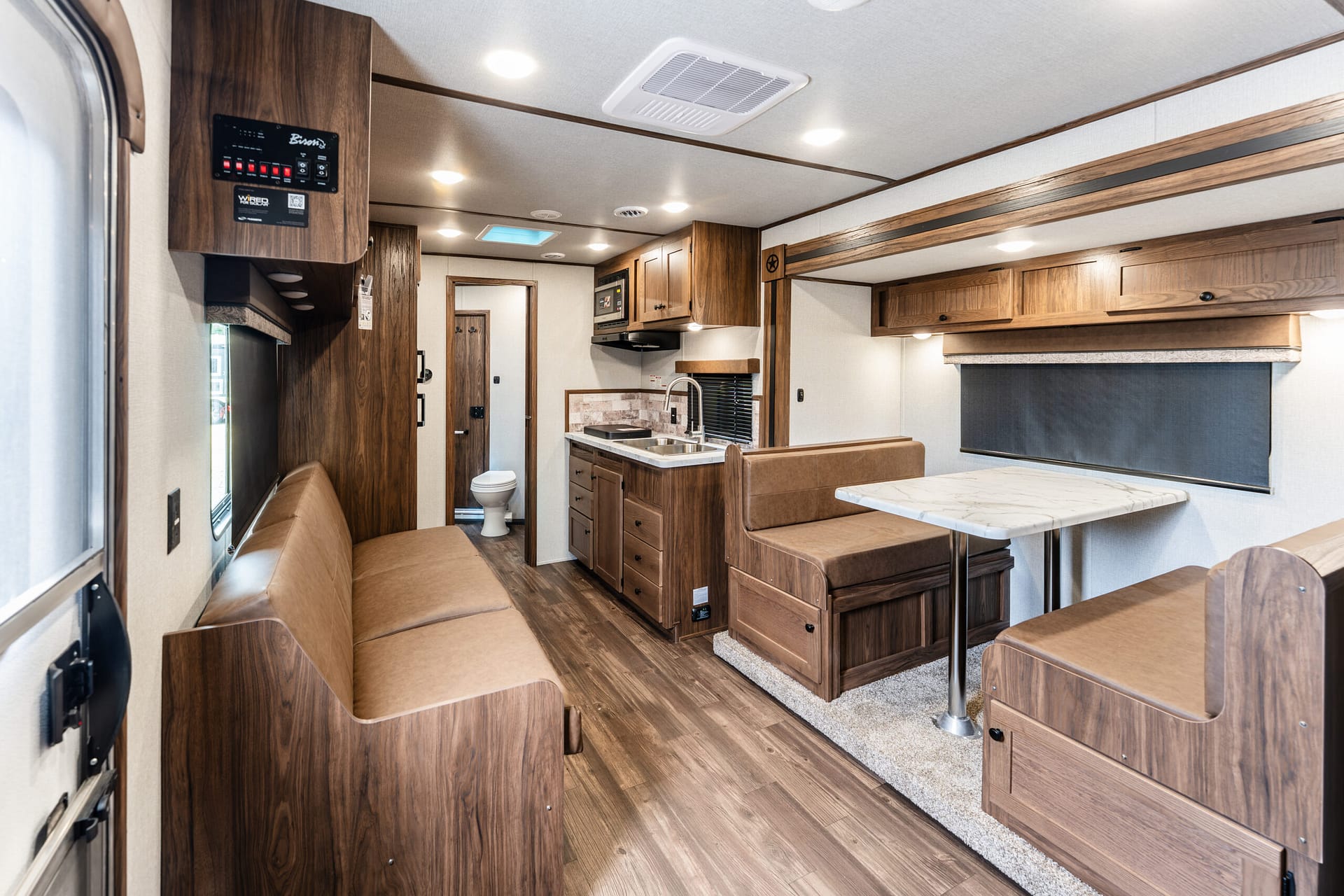 Interior of a wood-paneled RV living area with a brown leather L-shaped sofa, dining table, and a compact kitchen including sink and stove, overhead cabinets, and warm lighting.