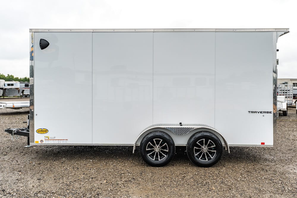 White enclosed cargo trailer with dual axles and diamond-plate fenders on a gravel lot.