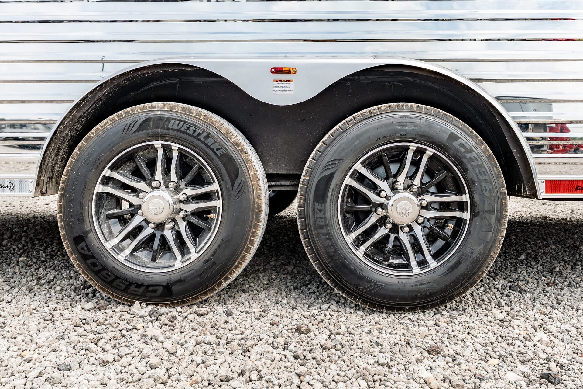Close-up of two trailer wheels with chrome rims and black fenders on a gravel surface.