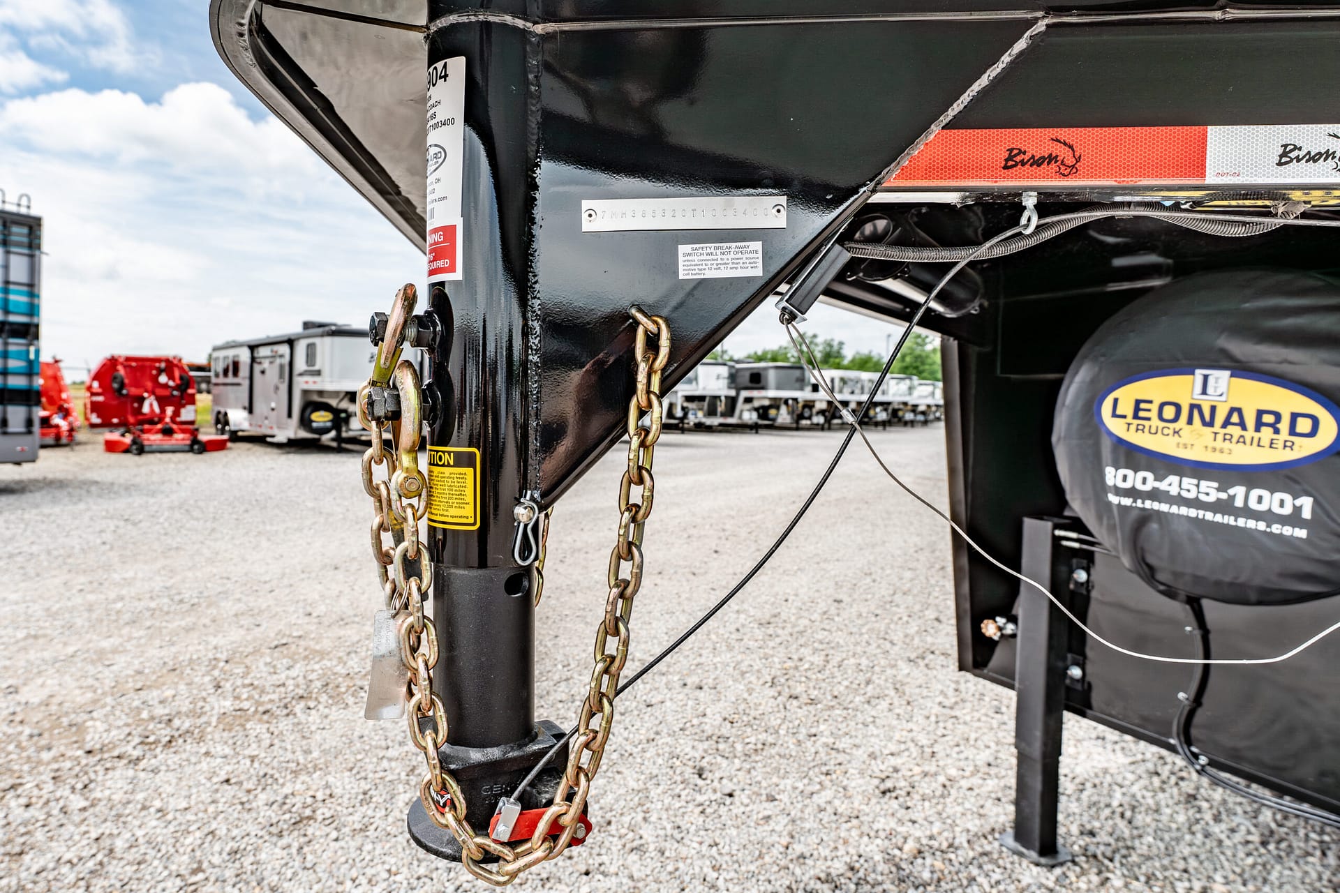 Close-up of a black trailer tongue with heavy safety chains and latch, Leonard trailer cover visible in the background in an outdoor lot.